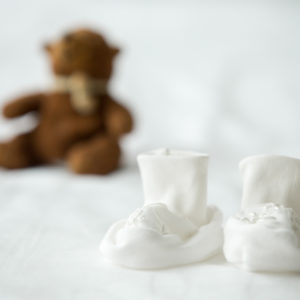 Close-up of white tiny booties for newborn baby on the bed. Cute teddy bear on the background. Maternity concept